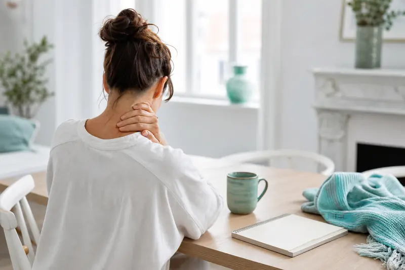 Personne massant sa nuque dans un intérieur lumineux, illustrant douleur et émotions.