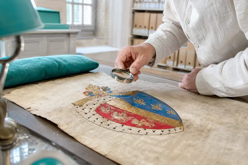 Mains examinant un blason héraldique peint sur parchemin avec une loupe.