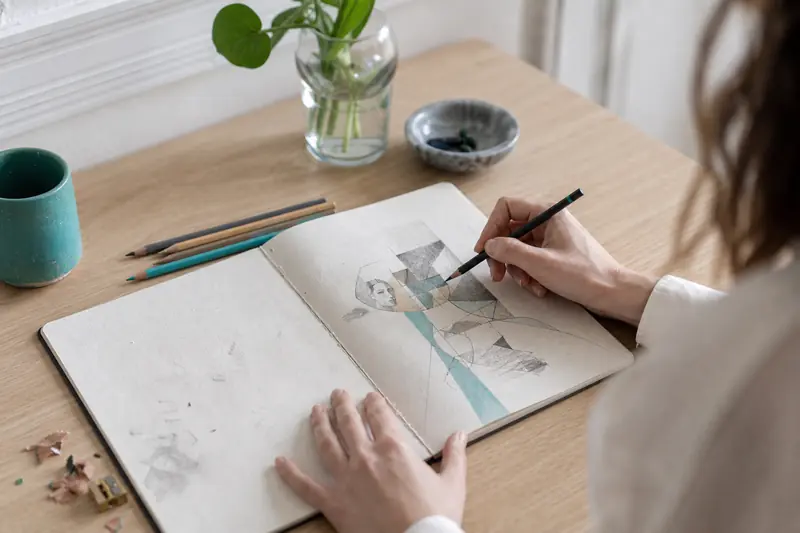 Carnet ouvert et mains traçant un dessin introspectif au crayon.
