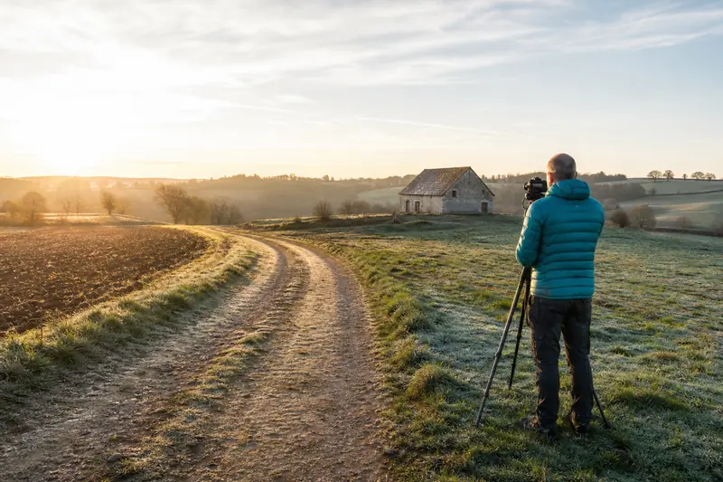 Photographe sur trépied face à un paysage rural de champs au lever du soleil